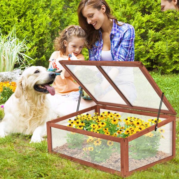 Portable cold frame with flowers inside and a lid lifted up that's set outside near a garden with a mom and daughter planting next to it and a white dog laying down next to them.