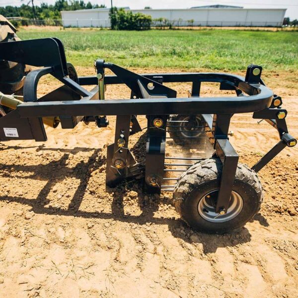 A potato digger digging up the ground in a field with building and a area of grass in the background.